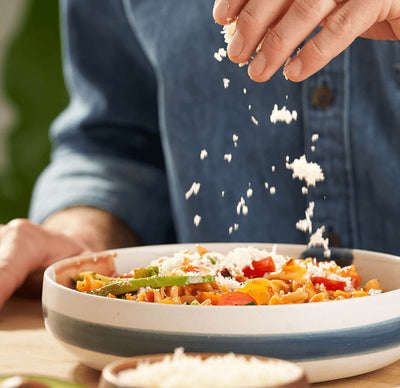 A close-up of a bowl of ZENB Rotini Pasta cooked with lightly-sauteed vegetables such as zucchini, carrots, and red bell pepper. A hand is above the bowl of pasta, garnishing the pasta with crumbled white cheese.